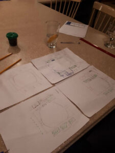 Table with sketches of circular diagrams on multiple sheets, pencils nearby, and a glass with lemon on a beige countertop.
