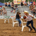 A woman in a white hat runs with a rope in a dirt rodeo arena, dodging plastic chairs as spectators watch from bleachers behind fencing.