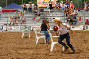 A woman in a white hat runs with a rope in a dirt rodeo arena, dodging plastic chairs as spectators watch from bleachers behind fencing.
