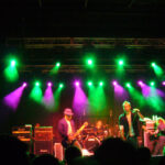 Live rock band on stage performing under green and purple lights, with amplifiers and drummer visible, audience silhouettes in the foreground.
