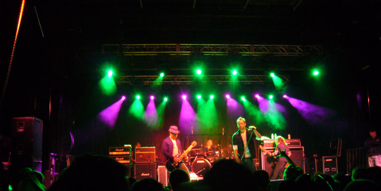 Live rock band on stage performing under green and purple lights, with amplifiers and drummer visible, audience silhouettes in the foreground.