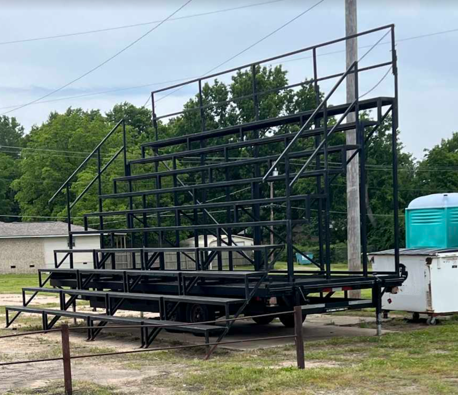 Stacked black metal bleachers on a trailer parked in a grassy lot, with trees and a portable toilet in the background.