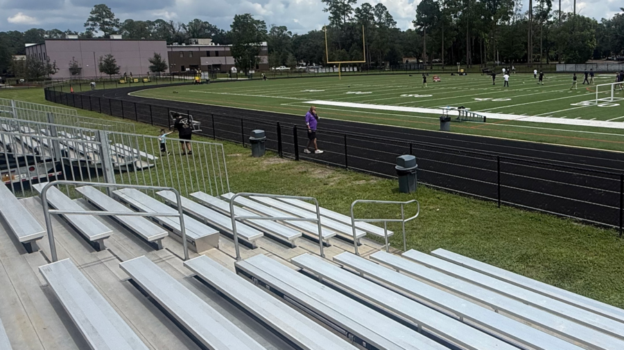 Empty metal bleachers overlook a green football field and running track with a few people near the sideline and goal posts in the distance.