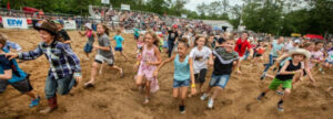 Children race across a dirt field at a crowded outdoor event with spectators in a bleacher stands backdrop.