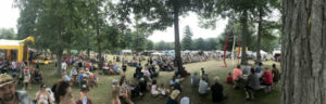 Crowd gathered at an outdoor festival in a park, with people sitting in groups under trees and colorful tents in the background.